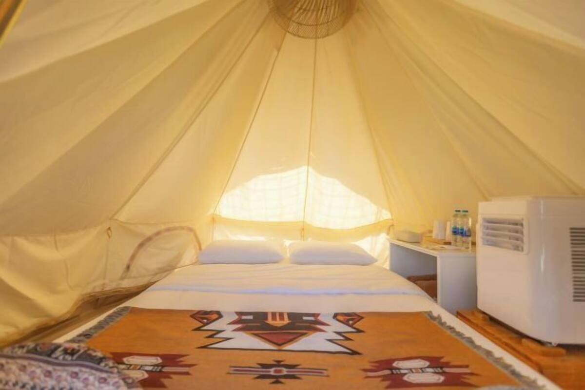Une pièce aménagée avec une tente, un lit, des oreillers, un tapis à motifs, une petite table avec quelques objets et un appareil électroménager blanc.