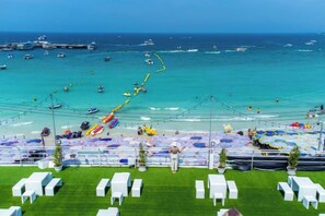 A beachside event with rows of white chairs and umbrellas, a grassy area, and a view of the ocean with boats.