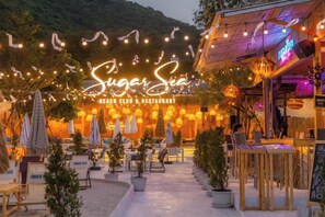 Un espace extérieur aménagé avec des tables et des parasols, une enseigne au néon indiquant « Sugar Sea Beach Club & Restaurant », et des plantes en pot.