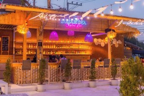 Un bar avec une enseigne au néon, des tables à l’extérieur et des plantes en pot.