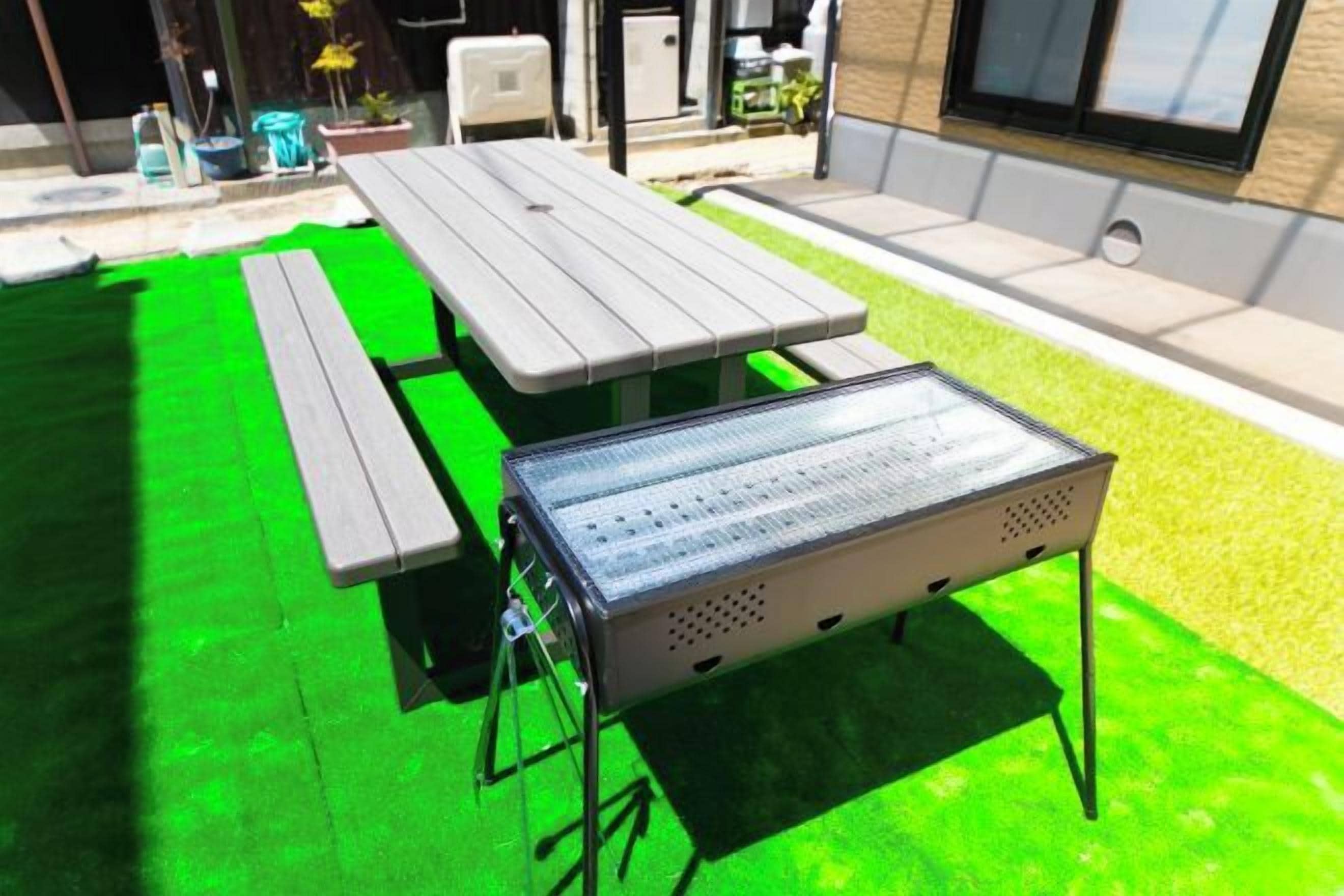 A picnic table with a metal grill and a bench, set on a green artificial turf.