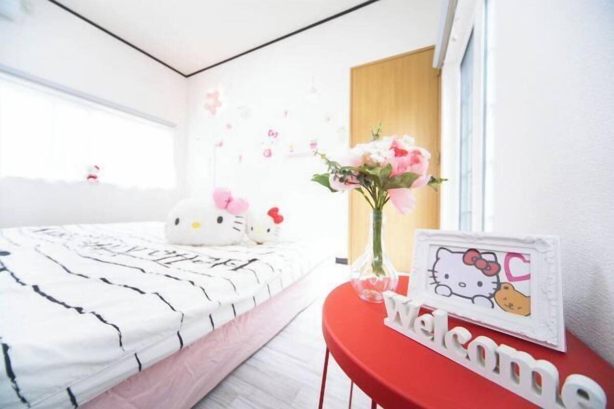 A bedroom with a bed, a red side table, a Hello Kitty sign, and a window.