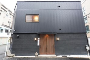 Un edificio moderno de dos pisos con fachada oscura, una puerta de madera y un ventanal pequeño.