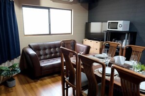 A living room with a brown leather sofa, wooden dining table, and chairs. A flat-screen TV is mounted on the wall above the sofa. - Japanese cottage Han (Osaka)