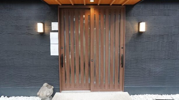 Entrada de um edifício moderno com porta de madeira, duas luzes fixadas na parede e uma parede com textura.