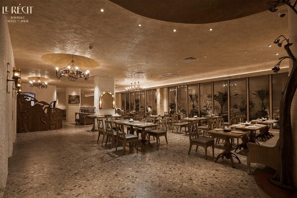 Une salle à manger spacieuse et élégamment aménagée, avec de nombreuses tables et chaises, des lustres et de grandes fenêtres offrant une vue sur l’intérieur.
