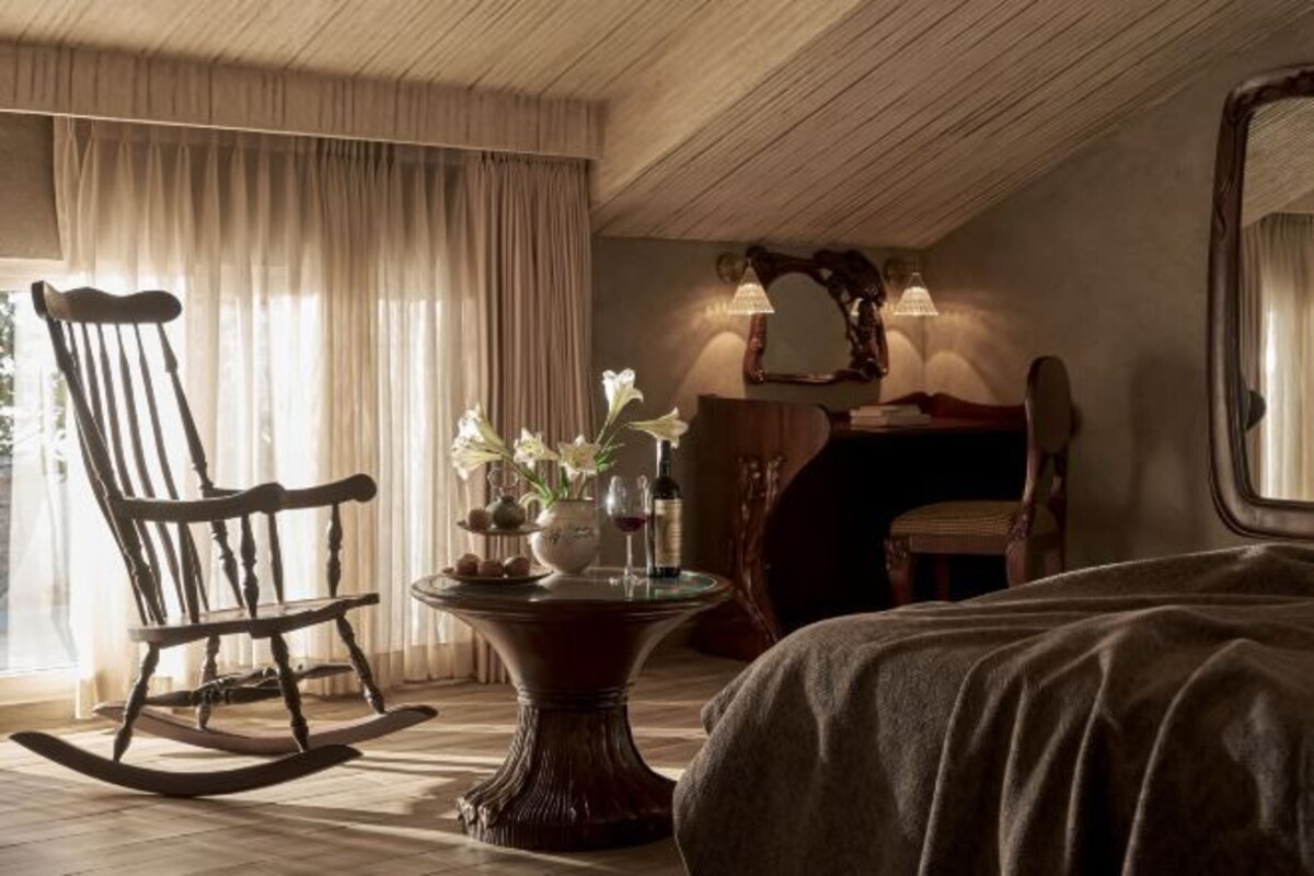 A cozy room with a wooden rocking chair, a small table with a vase of flowers, and a bed with a dark cover.
