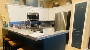A kitchen with white cabinets, a central island with stools, a microwave, and a refrigerator. - Chic Villa By The Bay- 48 TP (Laguna Vista)