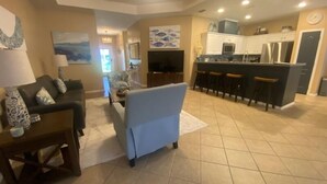 A spacious living area with a sofa, a coffee table, and a TV. There is a kitchen area with a bar and stools. - Chic Villa By The Bay- 48 TP (Laguna Vista)