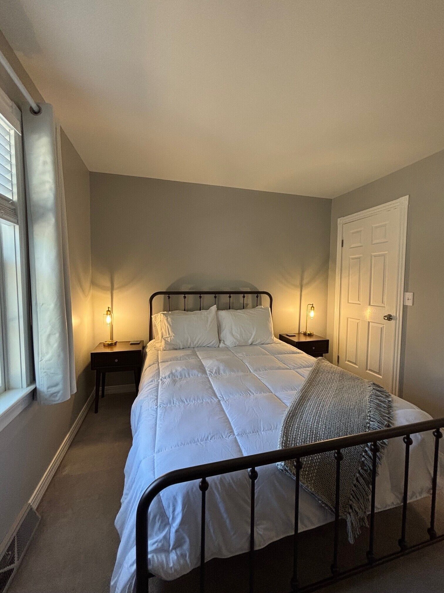 Un lit bien fait dans une pièce avec une fenêtre, deux tables de chevet avec des lampes et une porte.