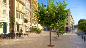 Una plaza pavimentada con mesas al aire libre, un árbol y edificios con balcones y ventanas.