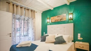 Una cama bien tendida con una colcha azul, almohadas blancas y un cabecero blanco. Hay una mesita de noche con una lámpara y una pequeña mesa junto a la cama.