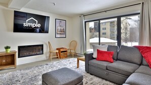 A modern living room with a grey sofa, a fireplace, a TV displaying 'simple vacation rentals', and a large window with a view of buildings.