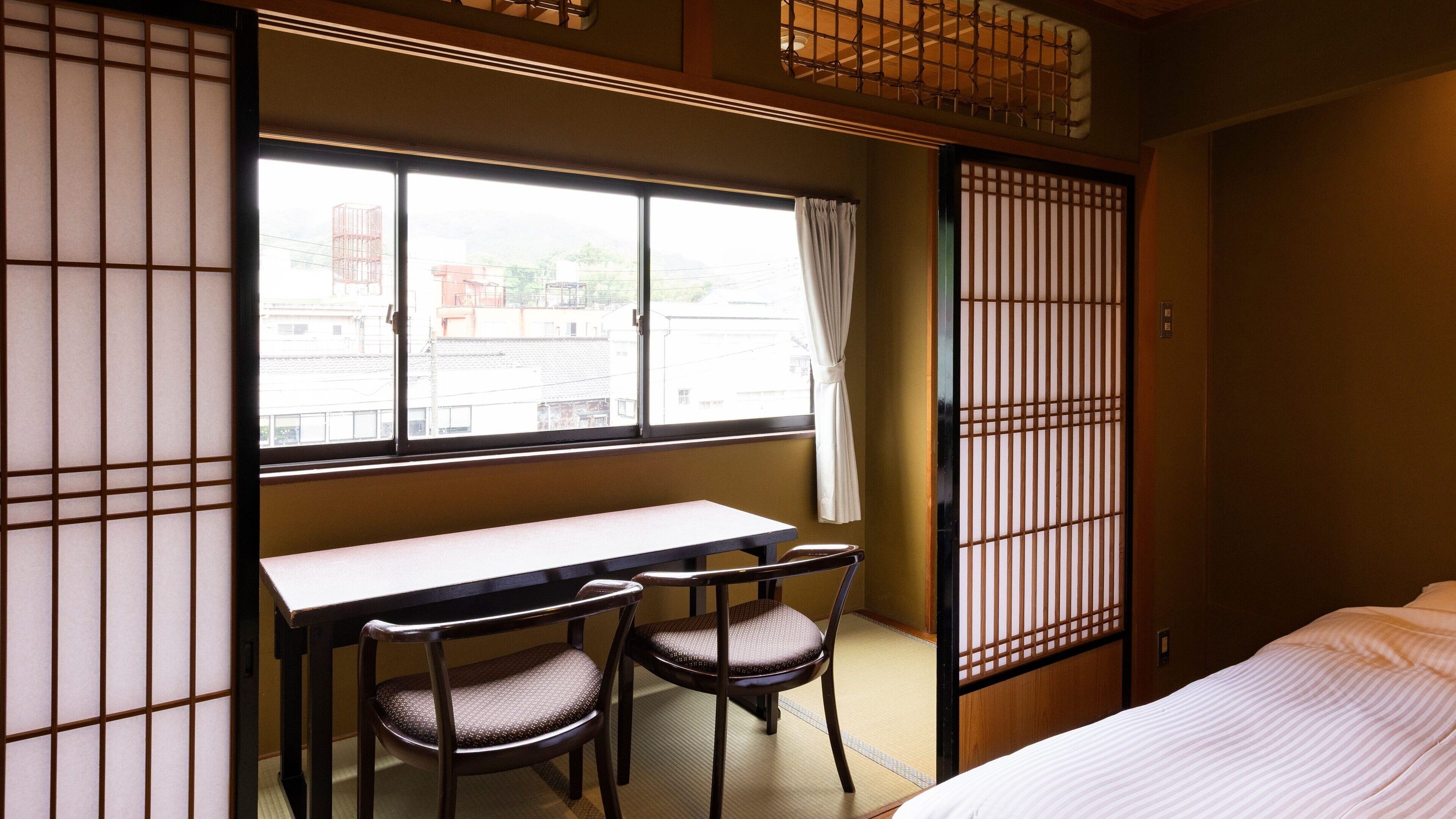 Une pièce comprenant un lit, un bureau, deux chaises, une fenêtre donnant sur des immeubles et une porte coulissante à motifs géométriques.
