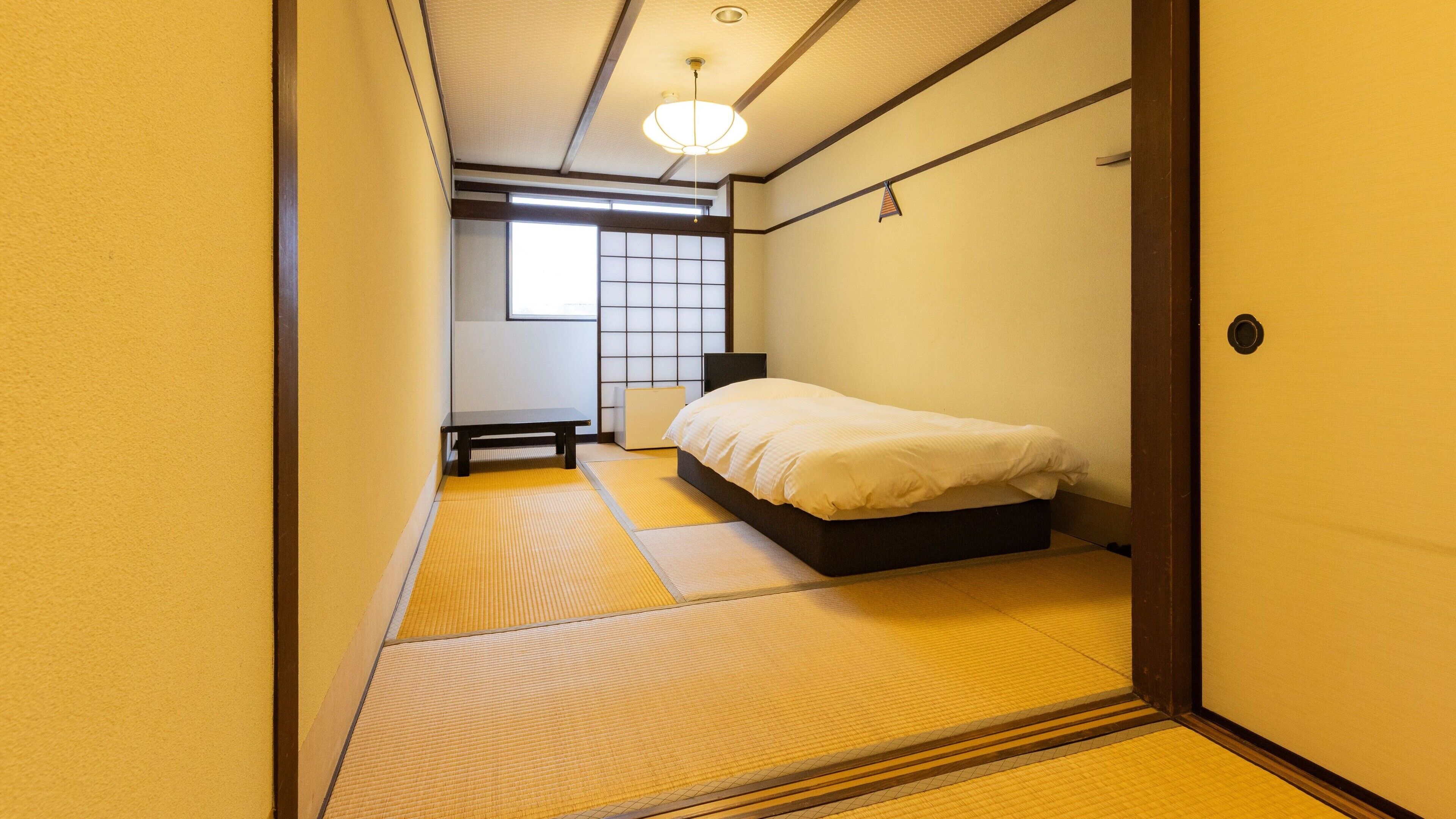 Une pièce de style japonais traditionnel, avec un sol recouvert de tatamis, un lit avec une literie blanche, une petite table et une porte coulissante.