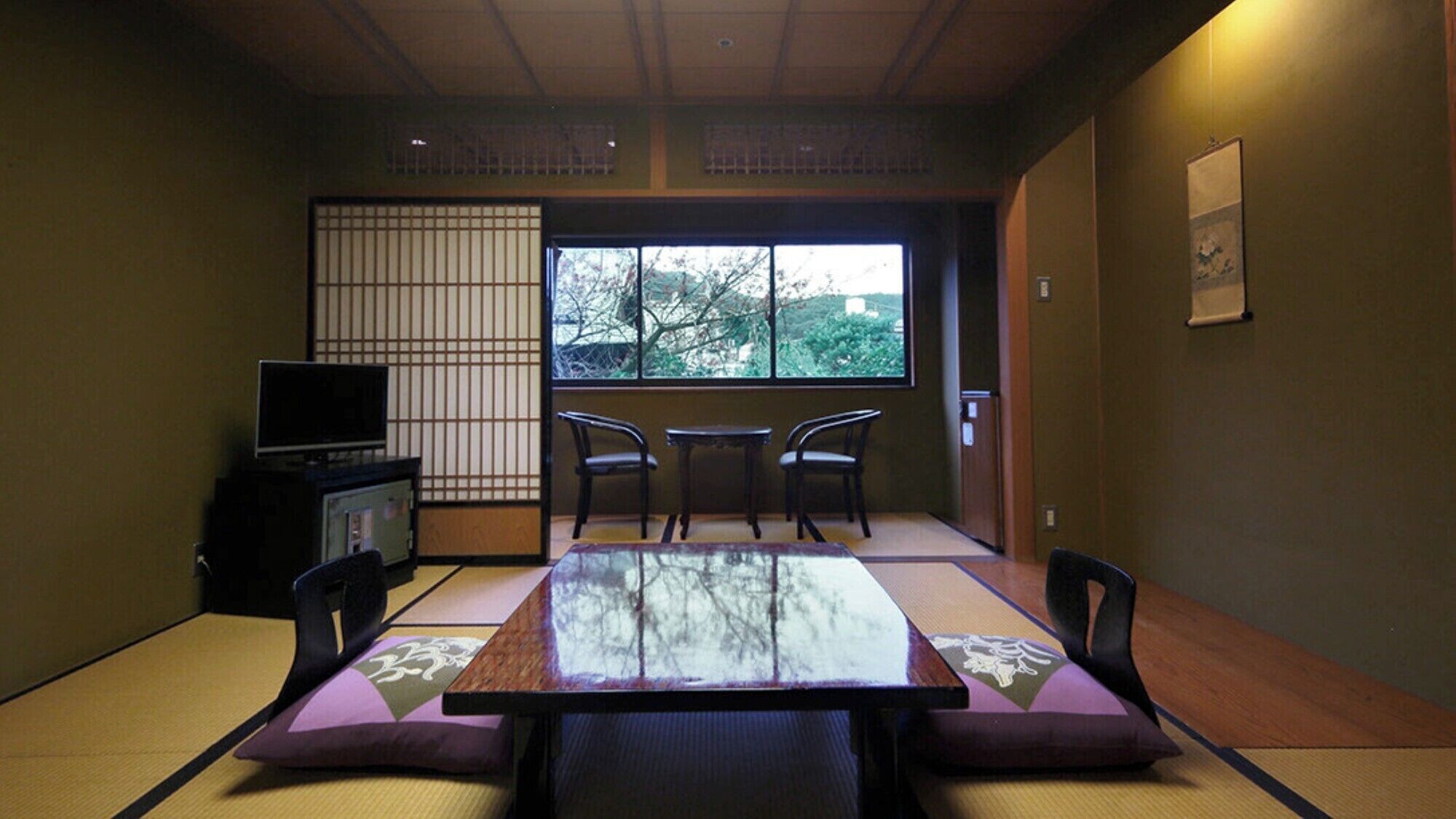 Une pièce de style japonais traditionnel, avec un sol recouvert de tatamis, une petite table basse avec des coussins, un téléviseur et une fenêtre donnant sur la verdure.