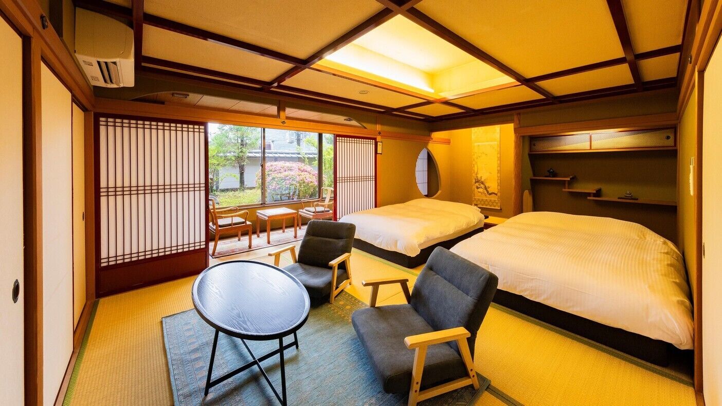 Une pièce de style japonais traditionnel, avec un sol recouvert de tatamis, deux lits, une petite table et une vue sur un jardin à travers des paravents en papier de riz (shoji).