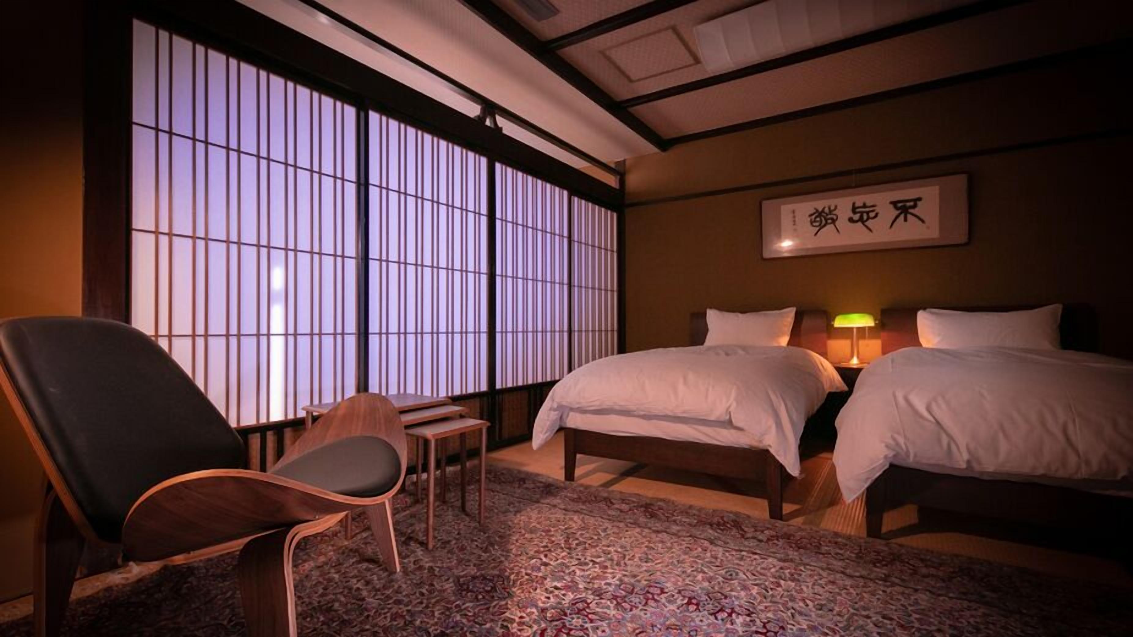 Une chambre d’hôtel avec deux lits, une chaise, une table et une grande fenêtre avec des stores verticaux.