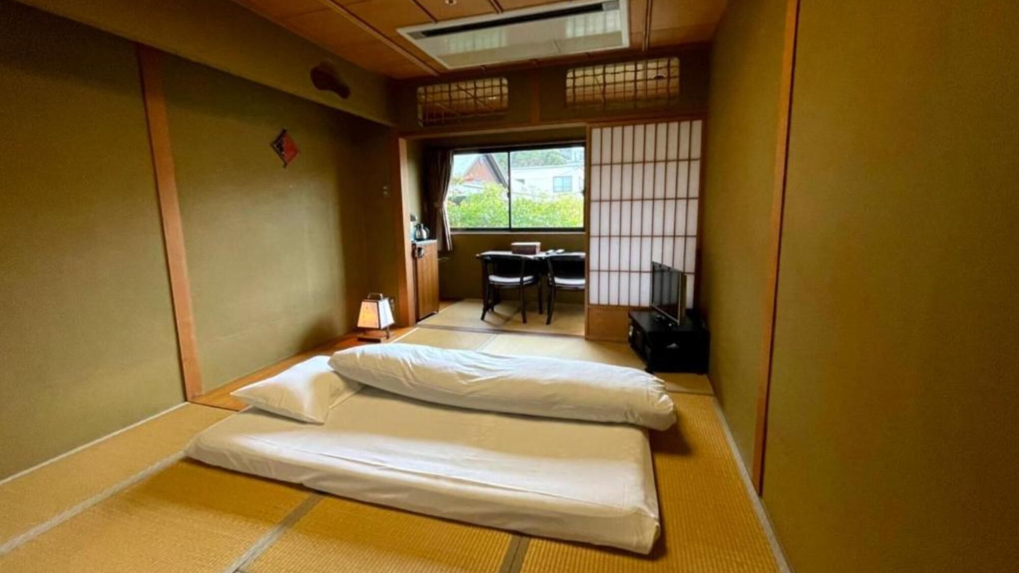 Une pièce de style japonais traditionnel, avec un sol recouvert de tatamis, un futon servant de lit, une petite table avec des chaises, un téléviseur et une fenêtre donnant sur un paysage verdoyant.
