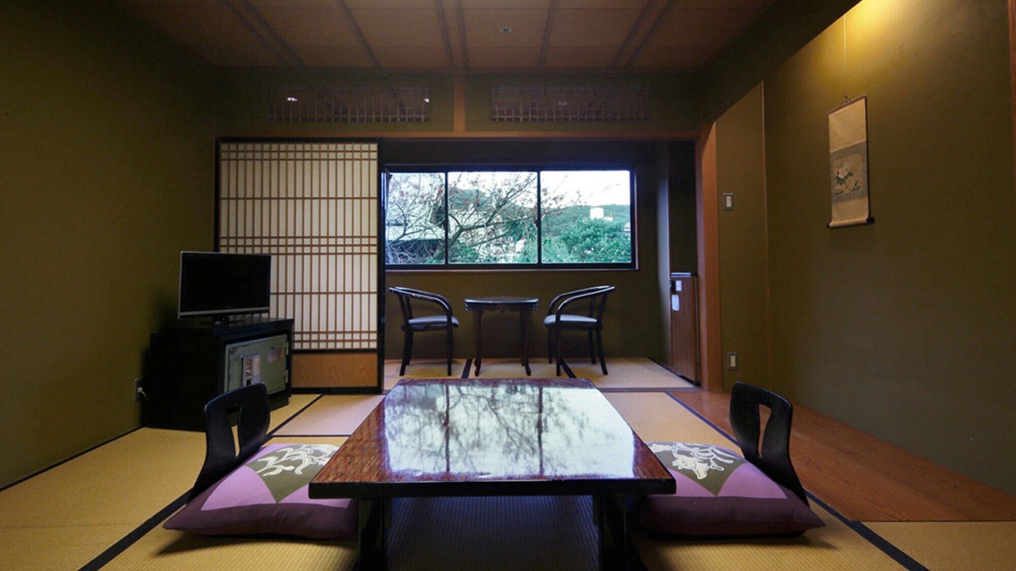 Une pièce de style japonais traditionnel, avec un sol recouvert de tatamis, une table basse avec des coussins, un téléviseur et une vue sur les arbres à travers un paravent en papier.