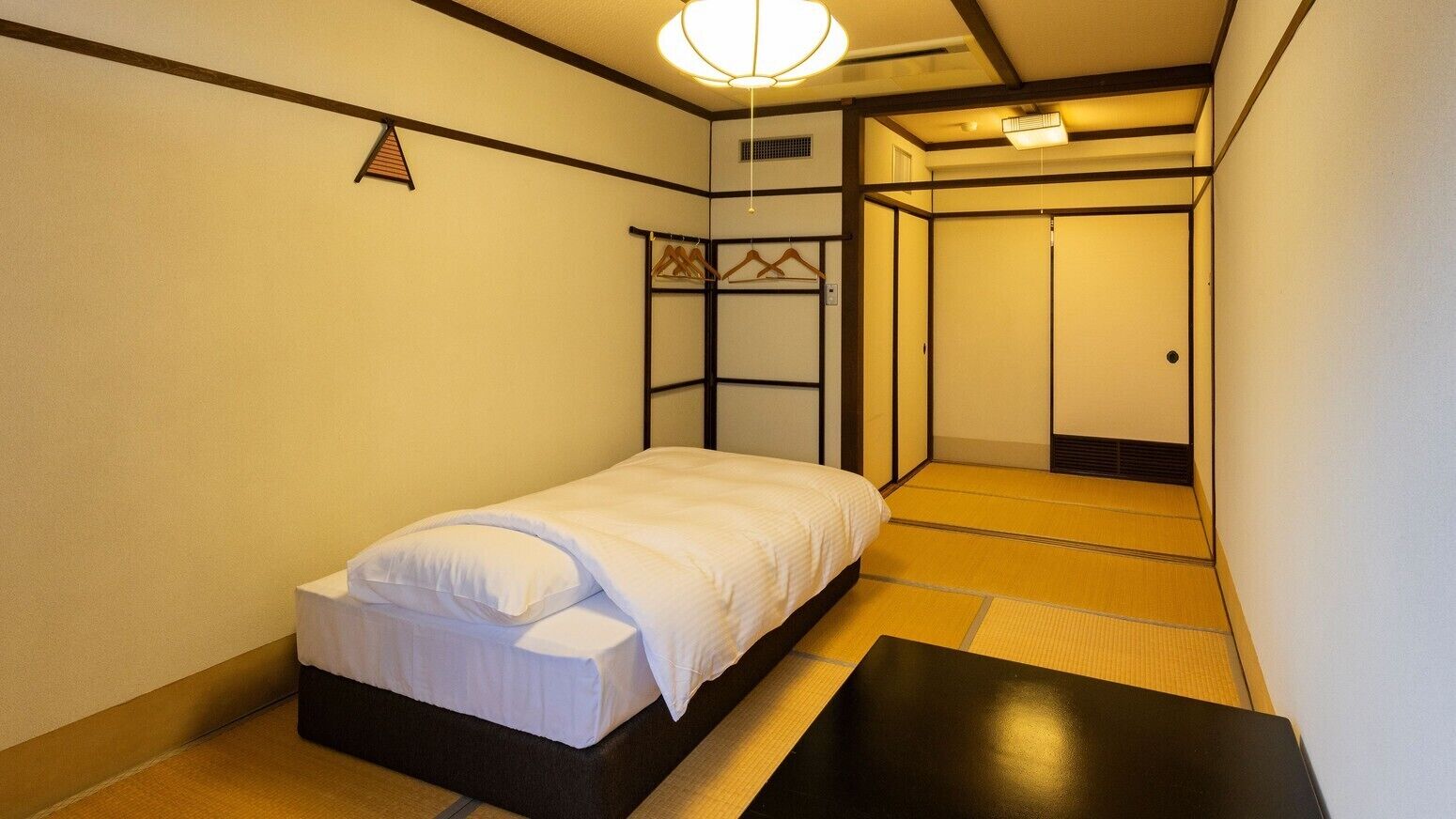 Une petite pièce, bien rangée, avec un lit, un banc, une armoire et un porte-vêtements.