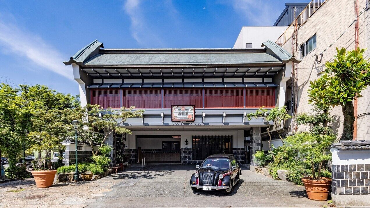 Un bâtiment traditionnel avec une voiture garée devant, une enseigne au-dessus de l’entrée et des plantes en pot tout autour.