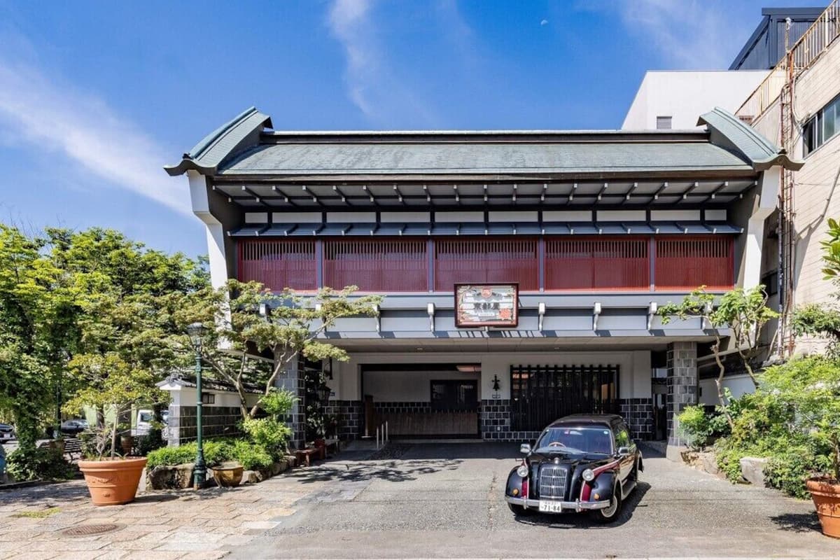 Un bâtiment traditionnel avec une voiture garée devant, une enseigne au-dessus de l’entrée et des plantes en pot tout autour.