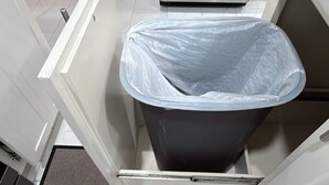 Una cocina con un refrigerador de acero inoxidable, un bote de basura gris y armarios blancos.