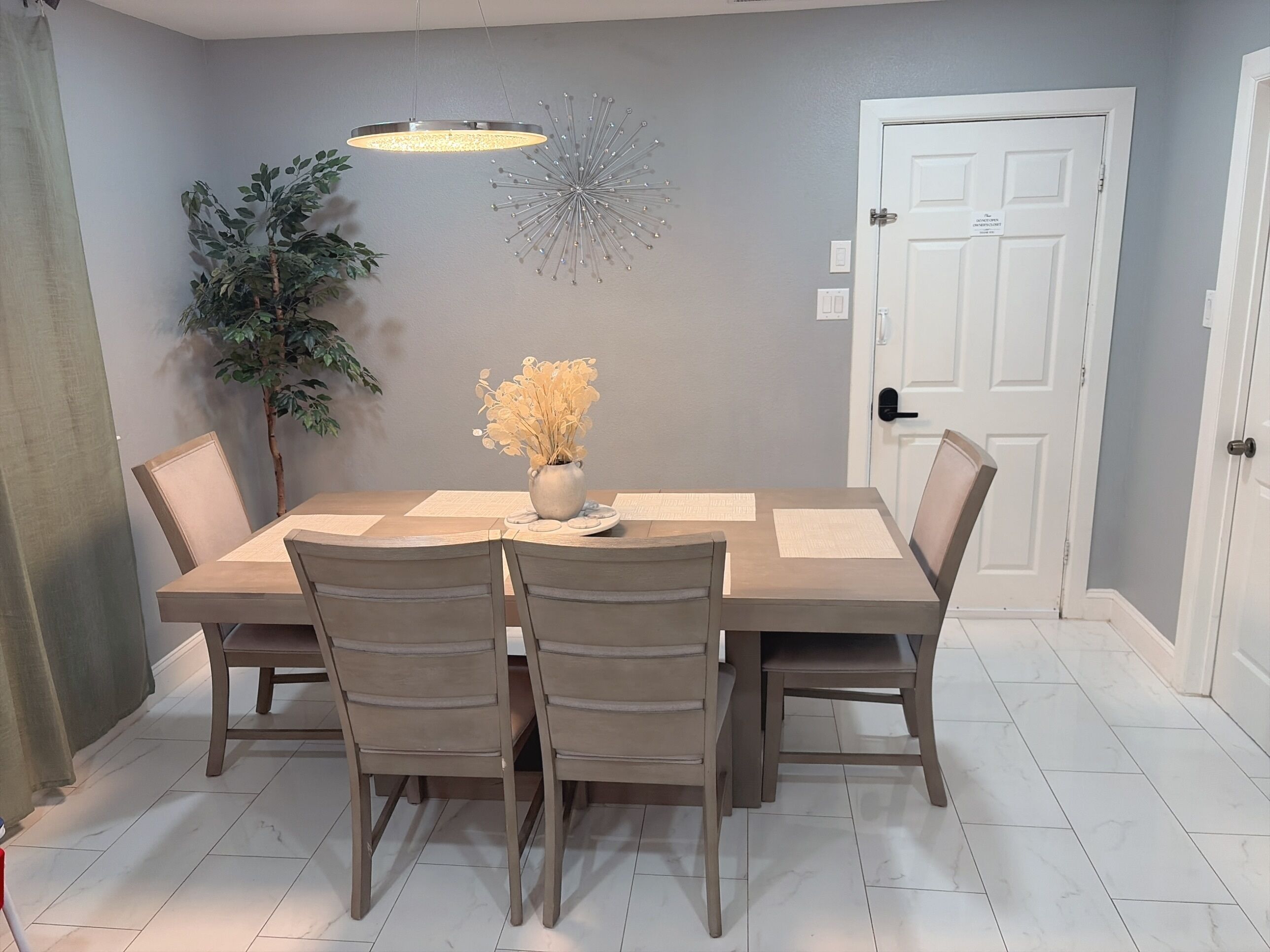 A dining area with a rectangular table, six chairs, a vase with flowers, a hanging light fixture, a wall decoration, and a door.