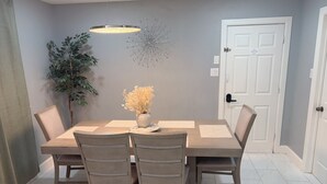 Un área de comedor con una mesa rectangular, seis sillas, un florero con flores, una lámpara colgante, un adorno en la pared y una puerta.