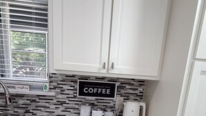 Una cocina con armarios blancos, encimera de mármol, estación de café y ventana con persianas.