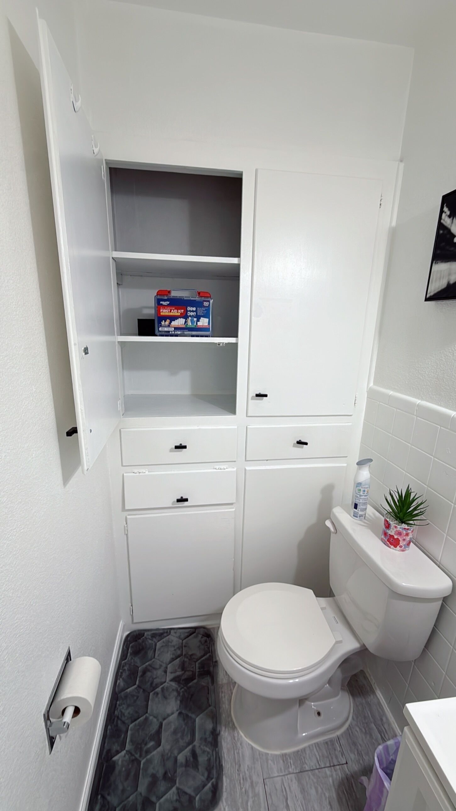 Ein kleines Badezimmer mit Toilette, einem Hängeschrank mit offenen Ablagen, einem Toilettenpapierhalter und einem kleinen Topf mit Pflanze.