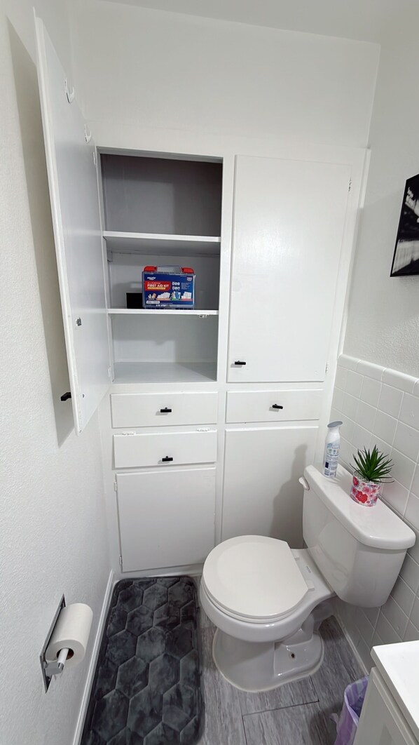 A compact bathroom with a toilet, a storage cabinet with open shelves, a toilet paper holder, and a small potted plant. - TEXANO INN Charming 3-bedroom house with WiFi and AC in welcoming Fort Worth (Fort Worth)
