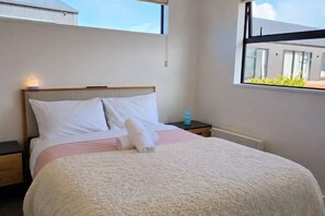 Ein ordentlich bezogenes Bett mit Kissen und Decke, ein Nachttisch mit einer Flasche sowie ein Fenster mit Blick auf Gebäude.