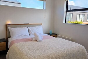 Un lit bien fait, avec des oreillers et une couverture, une table de chevet avec une bouteille, et une fenêtre donnant sur des immeubles.