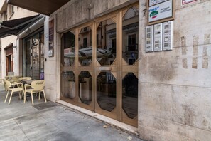 Un edificio con fachada de vidrio amplia, terraza con sillas de marca Coca-Cola y un letrero que indica bar de piano.