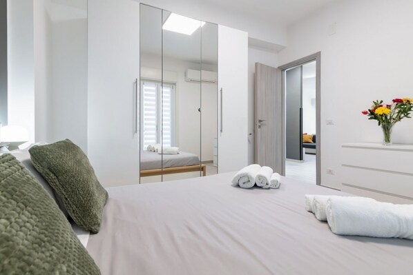 A modern hotel room with a large bed, a wardrobe with a mirror, a bedside table, and a vase with flowers.