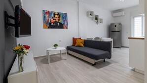 Ein modernes Wohnzimmer mit grauem Sofa, einem kleinen weißen Tisch, einem Gemälde an der Wand und einer Küchenzeile mit Kühlschrank.