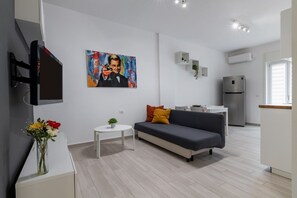 Una sala de estar moderna con un sofá gris, una mesita blanca, un cuadro en la pared y una zona de cocina con un refrigerador.