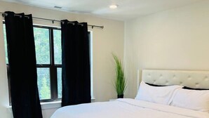 Ein modernes Schlafzimmer mit einem großen Bett, dunklem Holzboden, einem Fenster mit schwarzen Vorhängen und einer Topfpflanze.