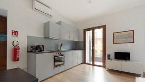 Une cuisine moderne avec des meubles gris, un four encastré, une machine à café et un téléviseur posé sur un meuble blanc.