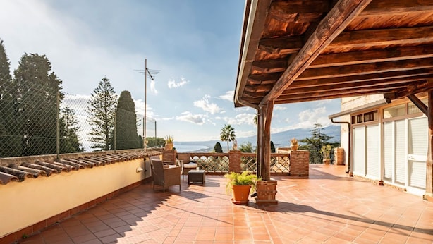 A rooftop terrace with tiled flooring, outdoor furniture, potted plants, and a view of the sea and mountains.