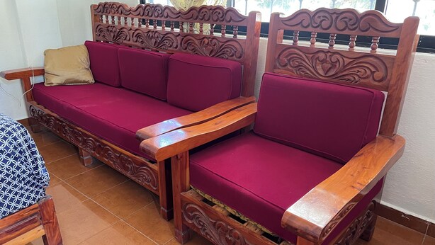 Um sofá de madeira com entalhes elaborados e encosto acolchoado, posicionado ao lado de uma mesa com toalha de padrão decorativo.