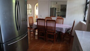 Uma área de refeições com uma mesa redonda, cadeiras de madeira, uma geladeira e uma cozinha ao fundo.