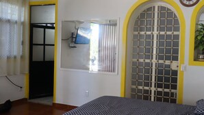 Um quarto com uma cama, uma janela com grades de metal, um relógio de parede e uma porta que dá para outro cômodo.
