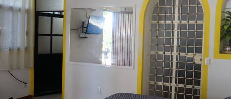 A room with a bed, a window with a metal grid, a wall clock, and a door leading to another room.