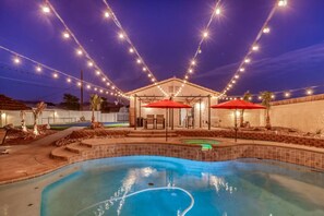 Een achtertuin met een zwembad, terras parasols, lichtjes en een overdekte zithoek.