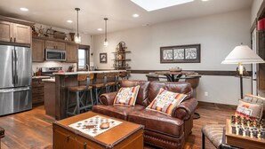 A spacious living room with a brown leather sofa, a wooden coffee table, and a chess set. A kitchen with stainless steel appliances and wooden cabinets is visible in the background. - Downtown Whitefish condo centrally located with great views and AC (Whitefish)