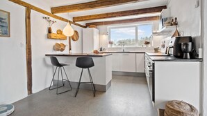 A modern kitchen with a wooden ceiling, a large window, and a built-in oven. There are two black chairs and a small island with a lamp. - Stunning apartment in Middelfart (Middelfart)