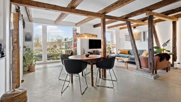 A spacious room with a high ceiling and exposed wooden beams, a round wooden table with chairs, a sofa, and a television mounted on the wall. - Stunning apartment in Middelfart (Middelfart)